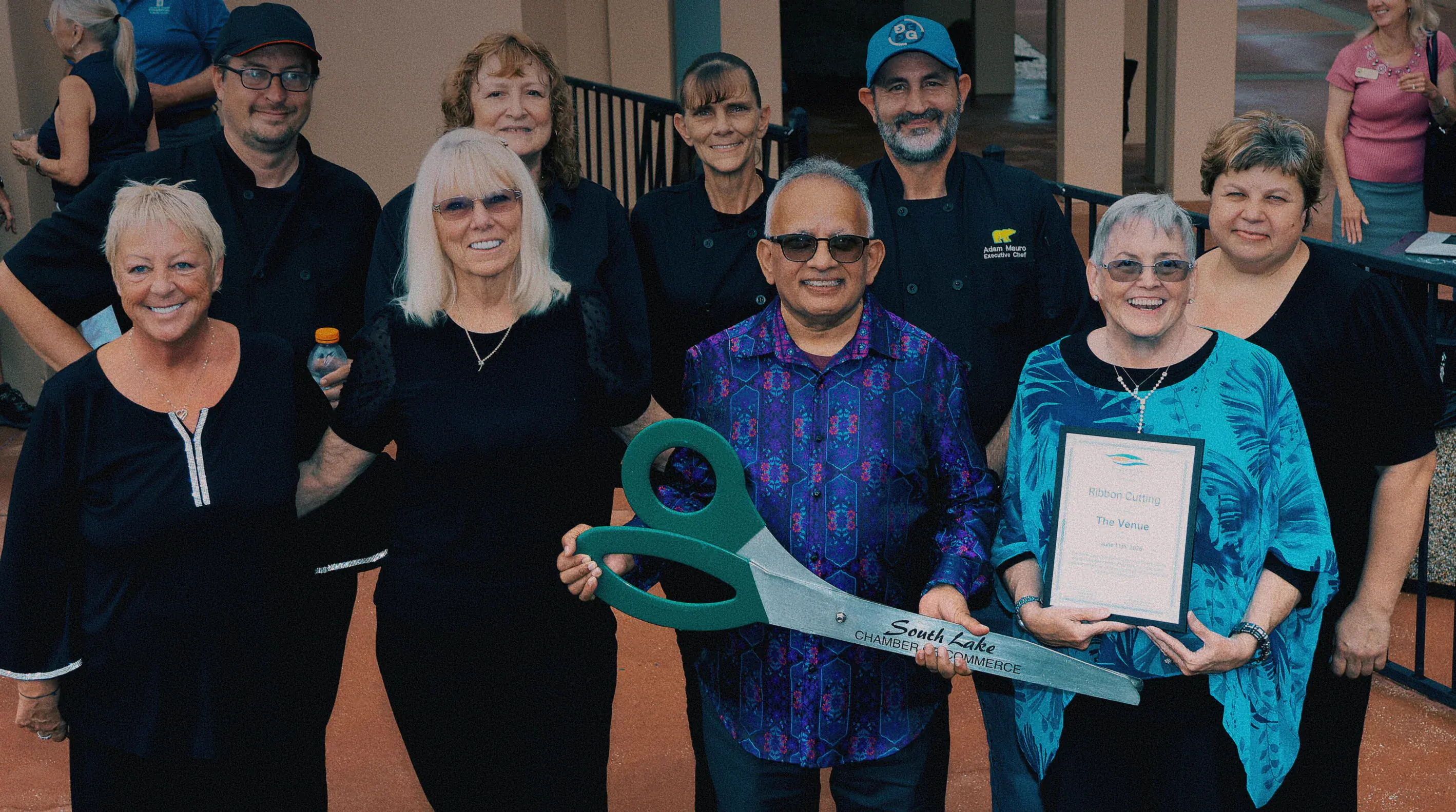 Ribbon cutting ceremony at The Venue Clermont grand opening with oversized ceremonial scissors, South Lake Chamber of Commerce plaque, and staff in chef coats